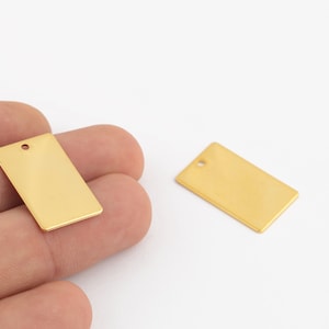 Puede incluir: Dos etiquetas rectangulares de metal de color dorado con un pequeño agujero en la parte superior. Las etiquetas están en blanco y listas para ser personalizadas.