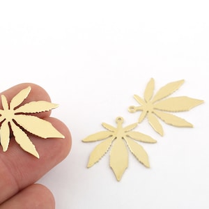 May include: Three gold-colored metal cannabis leaf charms with a hole at the top for hanging. The charms are on a white surface.