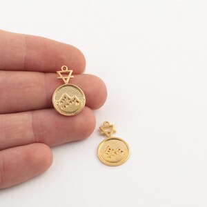 May include: Two gold-coloured pendant charms. Each charm features a circular design with a mountain range and a triangle symbol. The charms are small, with a loop at the top for attaching to a necklace or bracelet.