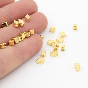 Puede incluir: Un grupo de cuentas cuadradas de metal dorado con un agujero en el centro. Las cuentas están esparcidas sobre una superficie blanca.