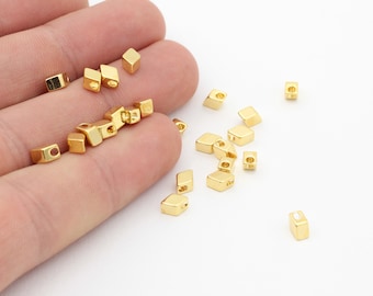 May include: A group of gold-colored metal square beads with a hole in the center. The beads are scattered on a white surface.