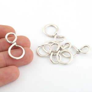 May include: Silver-toned metal jewellery findings in the shape of a figure eight. The findings are shown in a pile and individually in a hand.