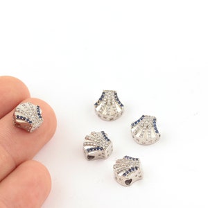 Puede incluir: Cinco cuentas de color plateado en forma de concha con cristales transparentes y azules. Las cuentas tienen un agujero en la parte superior para enhebrar.