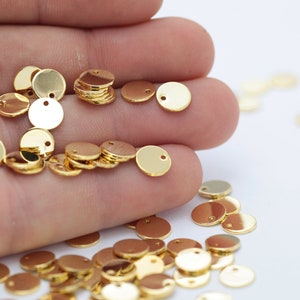 May include: A close-up of a hand holding a pile of gold-coloured metal discs with a small hole in the centre. The discs are scattered on a white surface.