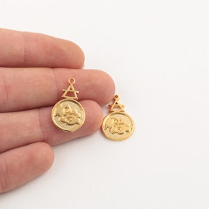 May include: Two gold-colored pendant charms. Each charm features a circular design with an embossed image and a triangular top with a small loop. The charms are likely for jewelry making.