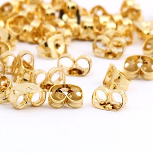 May include: A group of gold-colored earring backs, also known as earring stoppers, are scattered on a white surface. The earring backs are designed to secure earrings to the earlobe.