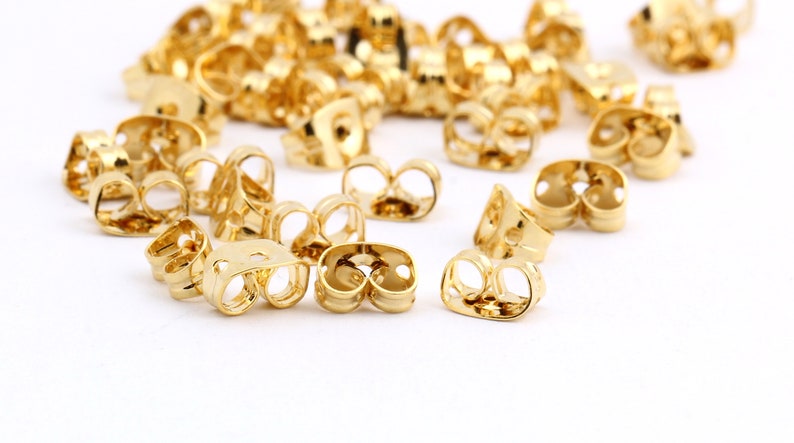 May include: A group of gold-colored earring backs, also known as earring stoppers, are scattered on a white surface. The earring backs are designed to secure earrings to the earlobe.