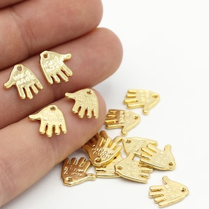 May include: A group of gold-colored metal hand charms with the words "Hand Made" engraved on them. The charms are scattered on a white surface and a hand is holding a few of them.