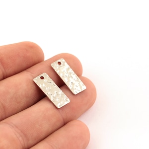 Puede incluir: Dos abalorios rectangulares de metal plateado con textura martillada y un pequeño agujero en la parte superior.