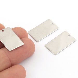 Pode incluir: Três etiquetas retangulares de metal prateado com um furo na parte superior. As etiquetas estão em branco e prontas para serem personalizadas.