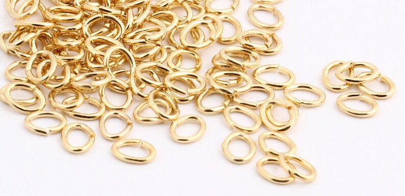 May include: A large pile of gold-coloured metal jump rings. The rings are oval shaped and are scattered on a white surface.
