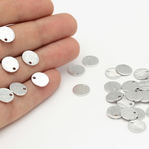 Könnte beinhalten: Ein Haufen runder silberner Metall-Anhänger mit einem Loch in der Mitte. Die Anhänger haben eine strukturierte Oberfläche.