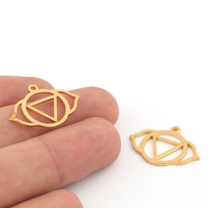 Pode incluir: Dois pingentes de metal dourado, cada um com um design de olho estilizado com um triângulo no centro. Os pingentes têm um pequeno laço na parte superior para prender a uma corrente ou outro componente de joia. O fundo é branco.