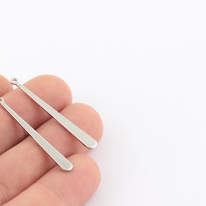 May include: Two silver metal earring pendants with a loop at the top and a pointed end. The pendants are approximately 5 cm long.