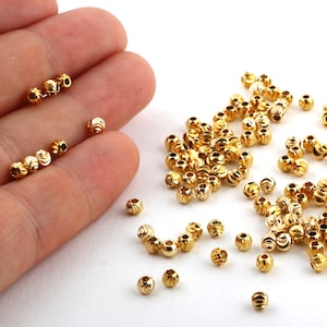 Puede incluir: Una gran cantidad de cuentas de metal de color dorado con una superficie texturizada y nervada. Las cuentas están dispersas sobre una superficie blanca.