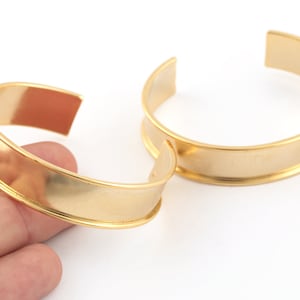Peut inclure: Deux bracelets manchette dorés. Les bracelets ont un design simple et ouvert avec une large bande plate. Le métal a une finition brillante. Un bracelet est tenu par une main, l'autre repose sur une surface blanche.
