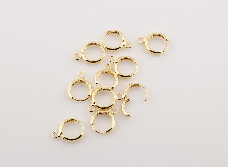 May include: A group of ten gold-colored earring hoops, all of the same size and style, are scattered on a white surface.
