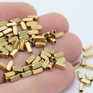 Könnte beinhalten: Nahaufnahme von kleinen, goldfarbenen Metallperlen. Jede Perle hat die Form eines rechteckigen Rohrs mit abgerundeten Enden. Die Perlen sind verstreut, was darauf hindeutet, dass sie lose sind und für die Schmuckherstellung oder Bastelarbeiten bestimmt sind.