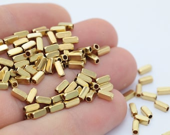 Könnte beinhalten: Nahaufnahme von kleinen, goldfarbenen Metallperlen. Jede Perle hat die Form eines rechteckigen Rohrs mit abgerundeten Enden. Die Perlen sind verstreut, was darauf hindeutet, dass sie lose sind und für die Schmuckherstellung oder Bastelarbeiten bestimmt sind.