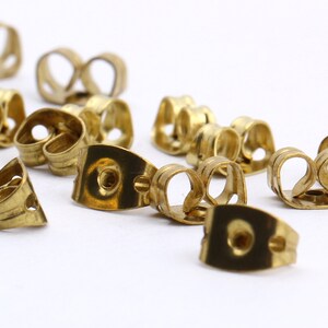 May include: A close-up of a group of gold-toned earring backs. The backs are made of metal and have a curved shape. They are designed to hold earrings in place.