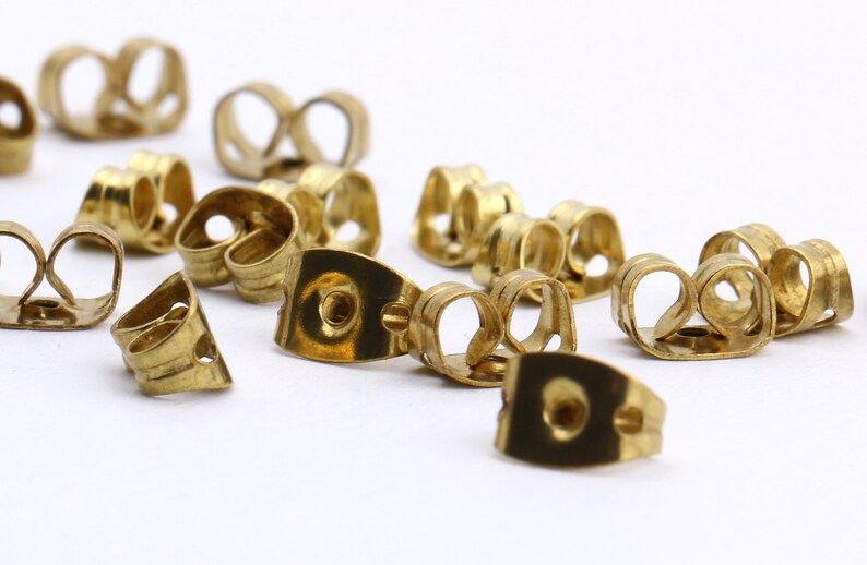 May include: A close-up of a group of gold-toned earring backs. The backs are made of metal and have a curved shape. They are designed to hold earrings in place.