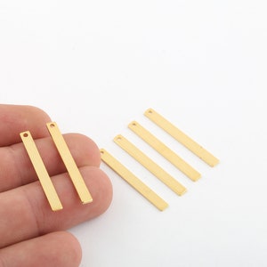 May include: Six gold-coloured metal bar pendants with a small hole at the top. The pendants are arranged in a staggered pattern on a white surface.
