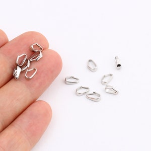 Pode incluir: Uma coleção de pequenos achados de joalheria prateados e brilhantes, incluindo formas ovais e retangulares abertas. Os achados estão espalhados em uma superfície branca, alguns sendo segurados em uma mão.