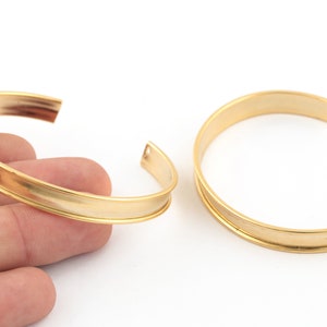 Puede incluir: Dos pulseras de metal doradas con una banda plana y ancha. Las pulseras están abiertas y listas para ser usadas.