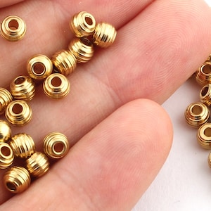 May include: A close-up of a hand holding a pile of gold-colored metal beads. The beads are round and have a textured surface. There are also a few beads scattered on a white surface.
