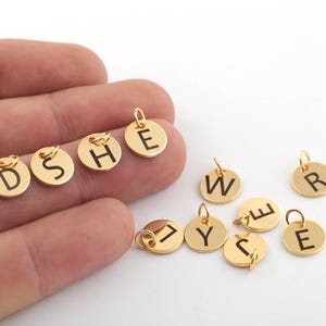 Może przedstawiać: Złote metalowe zawieszki z grawerowanymi czarnymi literami. Litery obejmują D, S, H, E, W, R, Y, C i E.