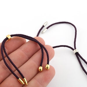 Puede incluir: Dos pulseras de cordón morado con detalles dorados y plateados. Una pulsera tiene una cuenta dorada en el extremo y la otra tiene una cuenta plateada en el extremo.