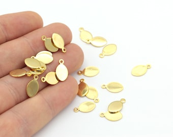 May include: A close-up of a hand holding a pile of gold oval-shaped metal charms with a loop at the top. The charms are scattered on a white surface.
