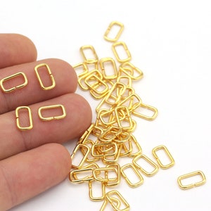 May include: A pile of gold-colored metal rectangular jump rings. The rings are small and have a smooth finish. They are commonly used in jewelry making and crafting.