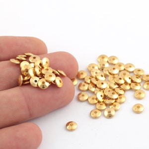 Könnte beinhalten: Ein Haufen goldfarbener Metallperlen, einige befinden sich in einer Hand, andere sind auf einer weißen Oberfläche verstreut.