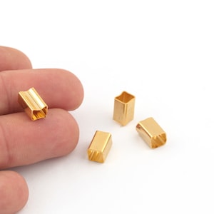 Pode incluir: Close-up de várias pequenas contas de metal retangulares douradas. As contas têm um design oco e aberto e são exibidas em um fundo branco. Uma conta é segurada pelos dedos de uma pessoa.