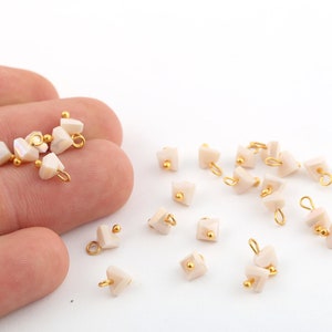 May include: Pale pink, geometric beads with gold-coloured loops and accents. The beads are scattered on a white surface, some held in a hand. These beads are likely for jewellery making.
