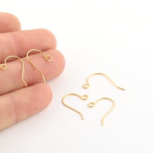 May include: Four gold-toned earring hooks, two are held in a hand, and two are lying on a white surface.