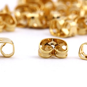 May include: Gold-toned earring backs, also known as earring stoppers, designed to secure earrings in place. The backs have a double-loop design with a small hole in the center.