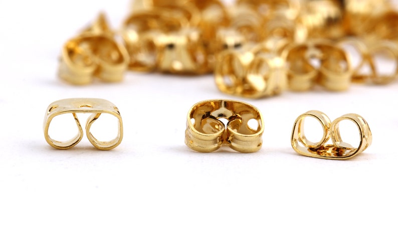 May include: Gold-toned earring backs, also known as earring stoppers, designed to secure earrings in place. The backs have a double-loop design with a small hole in the center.