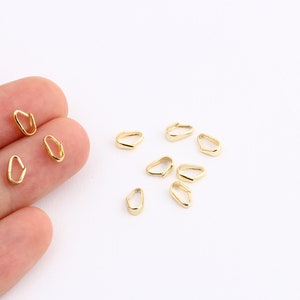 Puede incluir: Primer plano de baúles de colgantes de metal de color dorado. Los baúles son pequeños y de forma ovalada, con una ligera curva en la parte superior. Están dispuestos de forma dispersa sobre una superficie blanca.