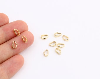Könnte beinhalten: Nahaufnahme von goldfarbenen Metall-Anhängerösen. Die Ösen sind klein und oval, mit einer leichten Biegung an der Oberseite. Sie sind auf einer weißen Oberfläche verstreut angeordnet.