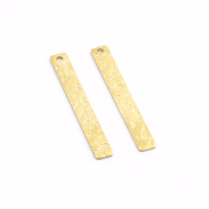 May include: Two gold-colored metal bar pendants with a textured surface. Each pendant has a small hole at the top for attaching a chain or string.