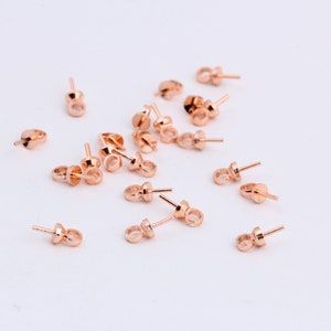 May include: A collection of rose gold earring posts and backs scattered on a white surface. The earring posts have a small, round loop at the bottom and a post extending from the top. The earring backs are flat with a small post.