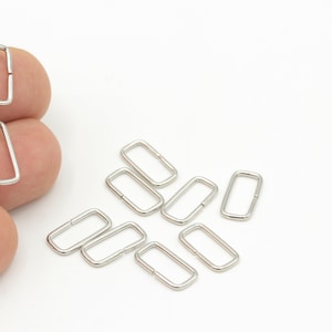 May include: A collection of silver-colored rectangular metal rings, some held in a hand, others scattered on a white surface. These small, open-ended rings are likely used for jewelry making or crafting projects. The rings are approximately 0.75 inches long.