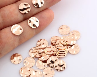 May include: A collection of 20 gold-toned metal round charms with a hammered texture and a hole in the centre. The charms are scattered on a white surface.