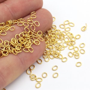 May include: A collection of small, gold-colored oval jump rings. The rings are scattered across a white surface, with some held in a hand. These jewelry-making components are ideal for crafting.