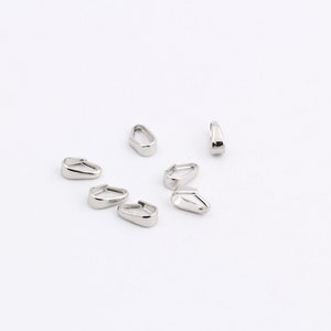 May include: A collection of shiny, silver-colored metal jewelry findings, possibly earring components, scattered on a white surface. The findings have a teardrop shape with a flat bottom and a curved top.