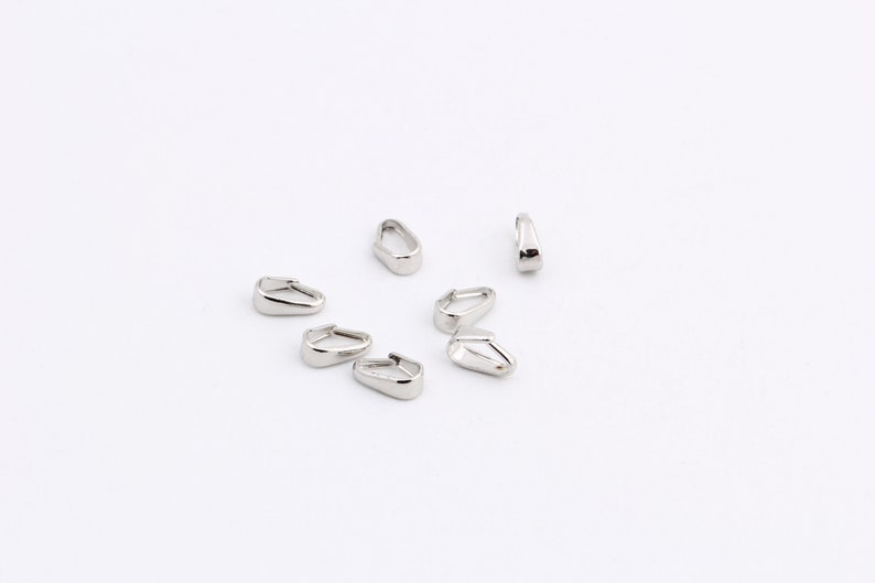 May include: A collection of shiny, silver-colored metal jewelry findings, possibly earring components, scattered on a white surface. The findings have a teardrop shape with a flat bottom and a curved top.