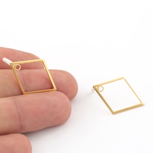 May include: A pair of gold-toned earrings with a square shape and a small circle at the top. The earrings are shown on a hand.