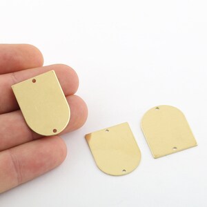 Op de afbeelding: Drie goudkleurige metalen labels met afgeronde randen en twee gaten elk. De labels hebben de vorm van een afgeronde rechthoek.